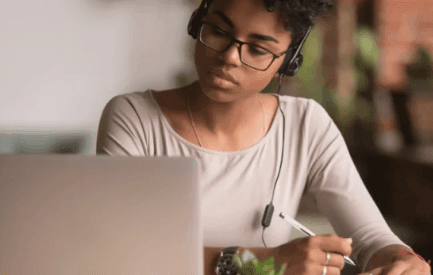 Woman wearing glasses and headphones working on a laptop, writing notes in a notebook.